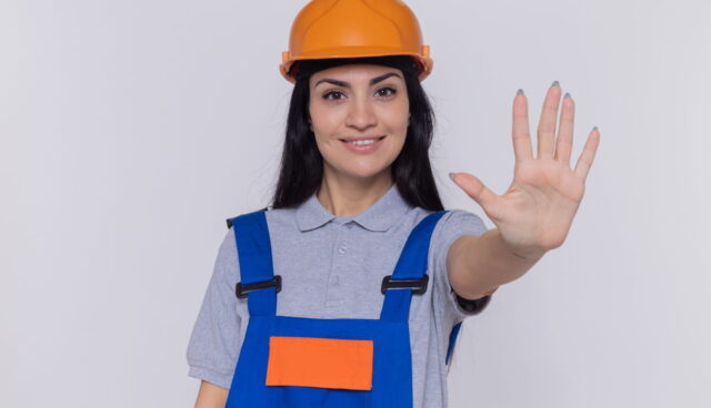 mulher jovem construtora em uniforme de construcao e capacete de seguranca olhando para frente sorrindo confiante fazendo gesto de parada com a mao aberta em pe sobre a parede branca
